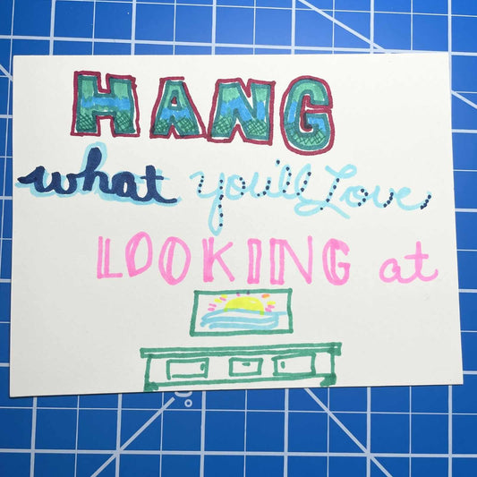 A hand-drawn card on a blue cutting mat featuring colorful lettering that says "HANG what you'll Love LOOKING at," emphasizing the importance of choosing a meaningful piece of art for your home.