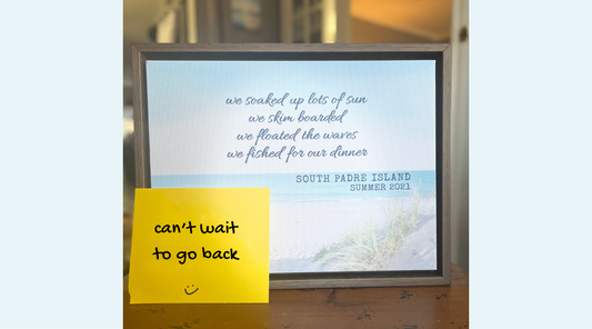 A framed beach art print sits on a wooden surface, with a yellow sticky note reading “can’t wait to go back” placed on the lower left corner.