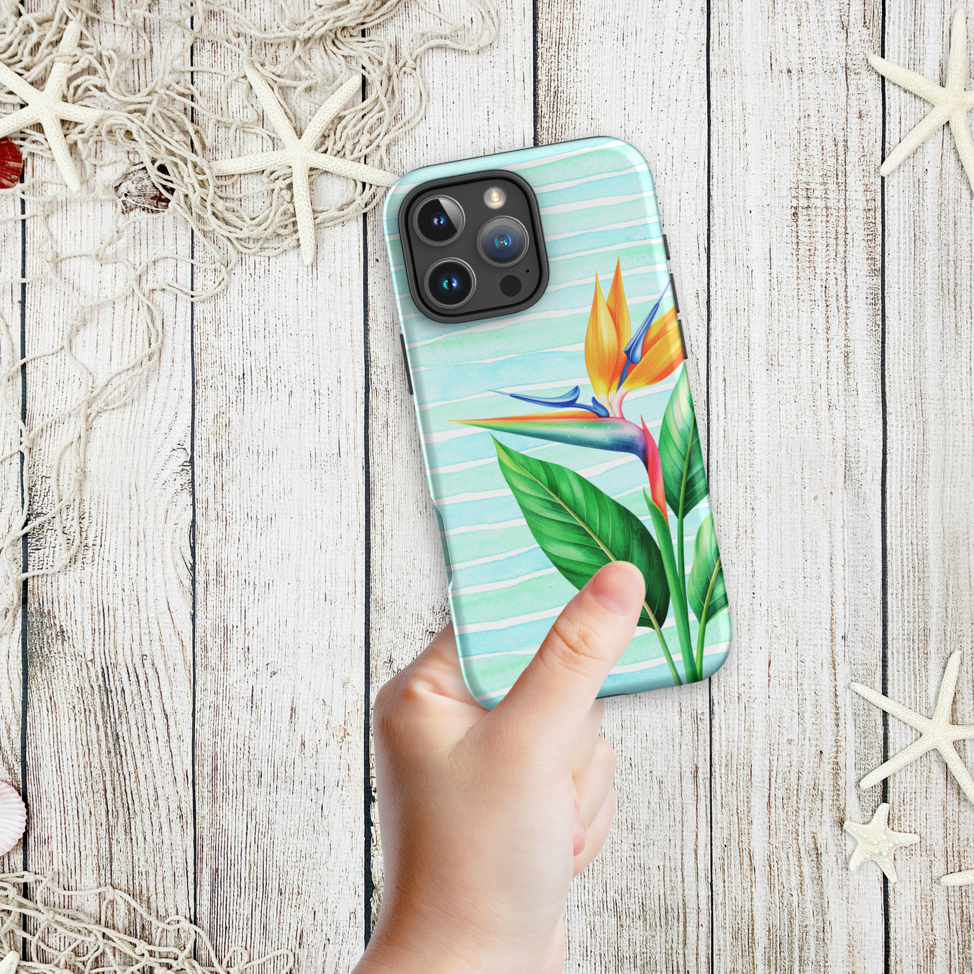 Hand holding a coastal phone case iPhone design with a tropical bird of paradise flower on aqua stripes, shown against a wood and starfish background.