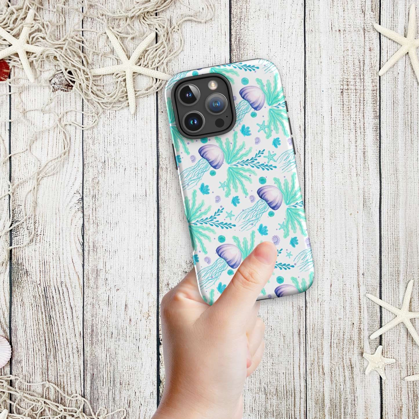 3. Hand holding a coastal phone case iPhone design with aqua and lavender jellyfish floating among seaweed, shown against a beach-inspired background with starfish.