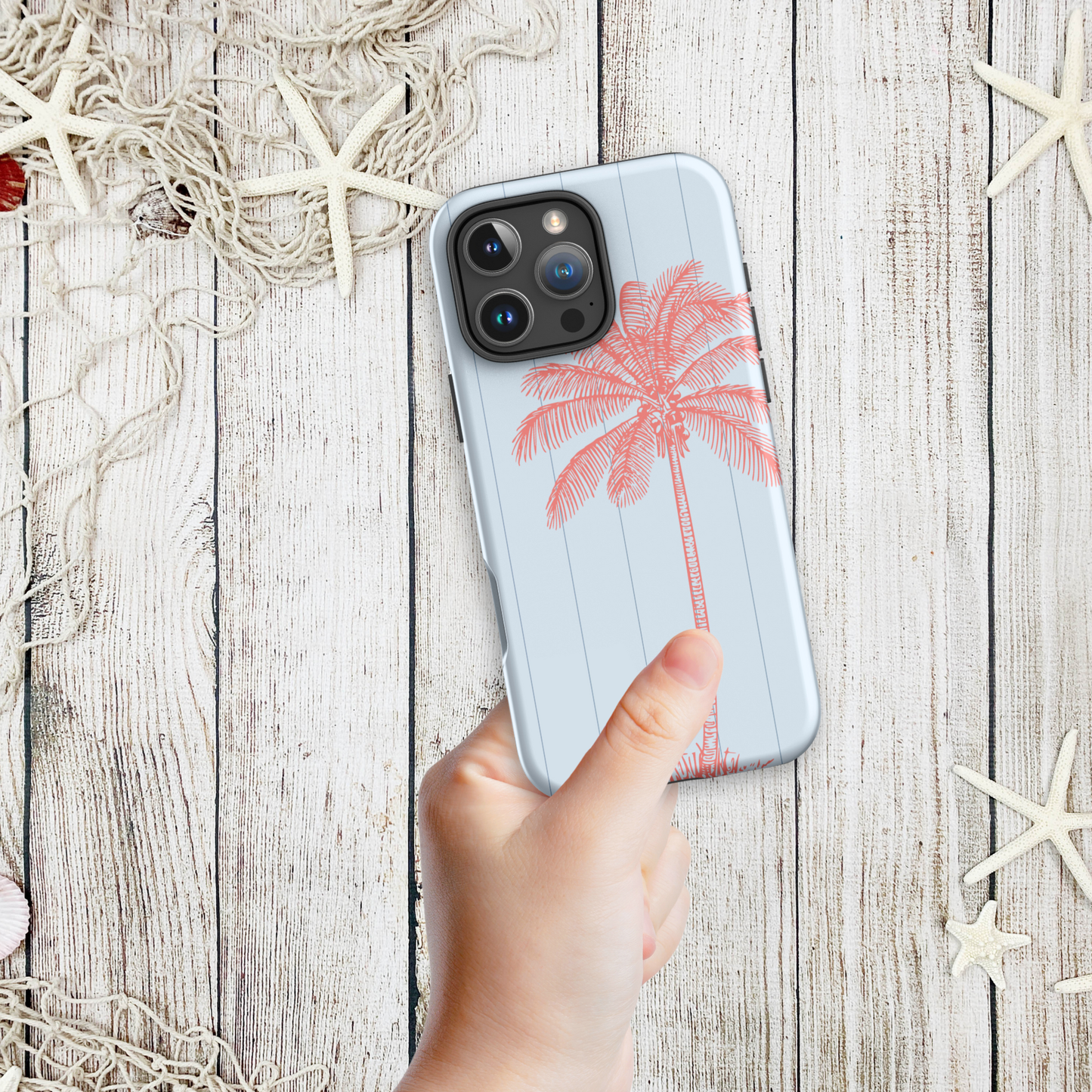 Hand holding a coastal iPhone case with a single pink palm tree sketch against a pale blue background, styled with starfish and nautical netting.