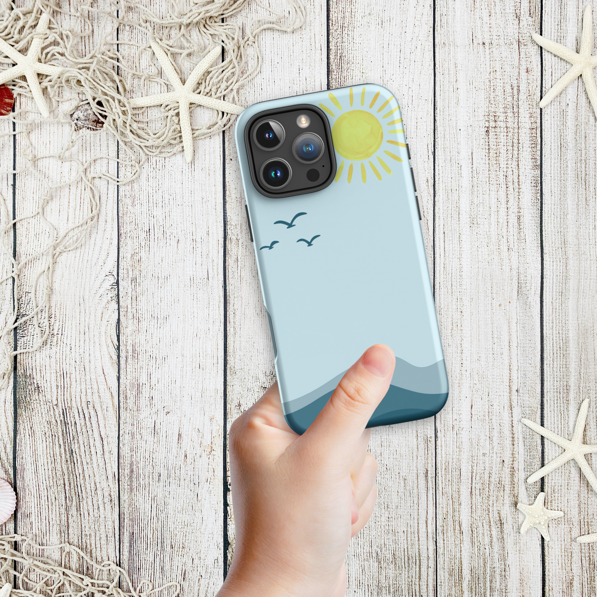 Hand holding a coastal phone case iPhone design with a minimalist ocean horizon, yellow sun, and flying seabirds, styled on a weathered wood background with starfish.
