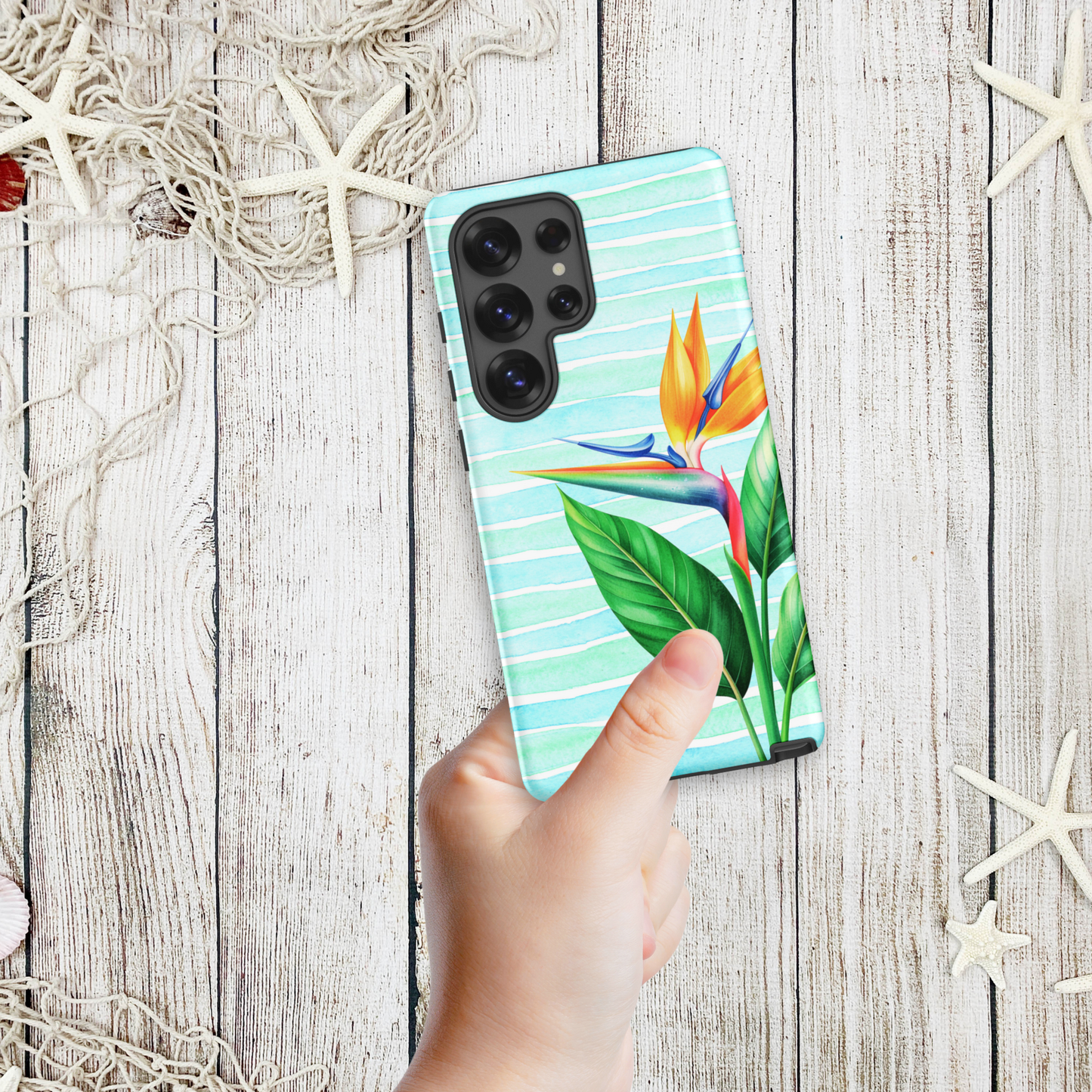 Hand holding a coastal Samsung phone case with a tropical bird of paradise flower on aqua stripes, shown against a wood and starfish background.