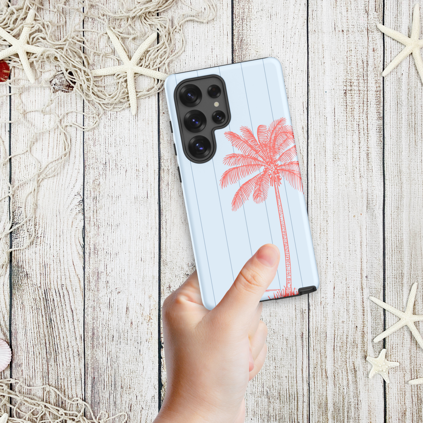 Hand holding a coastal Samsung phone case with a single pink palm tree sketch against a pale blue background, styled with starfish and nautical netting.