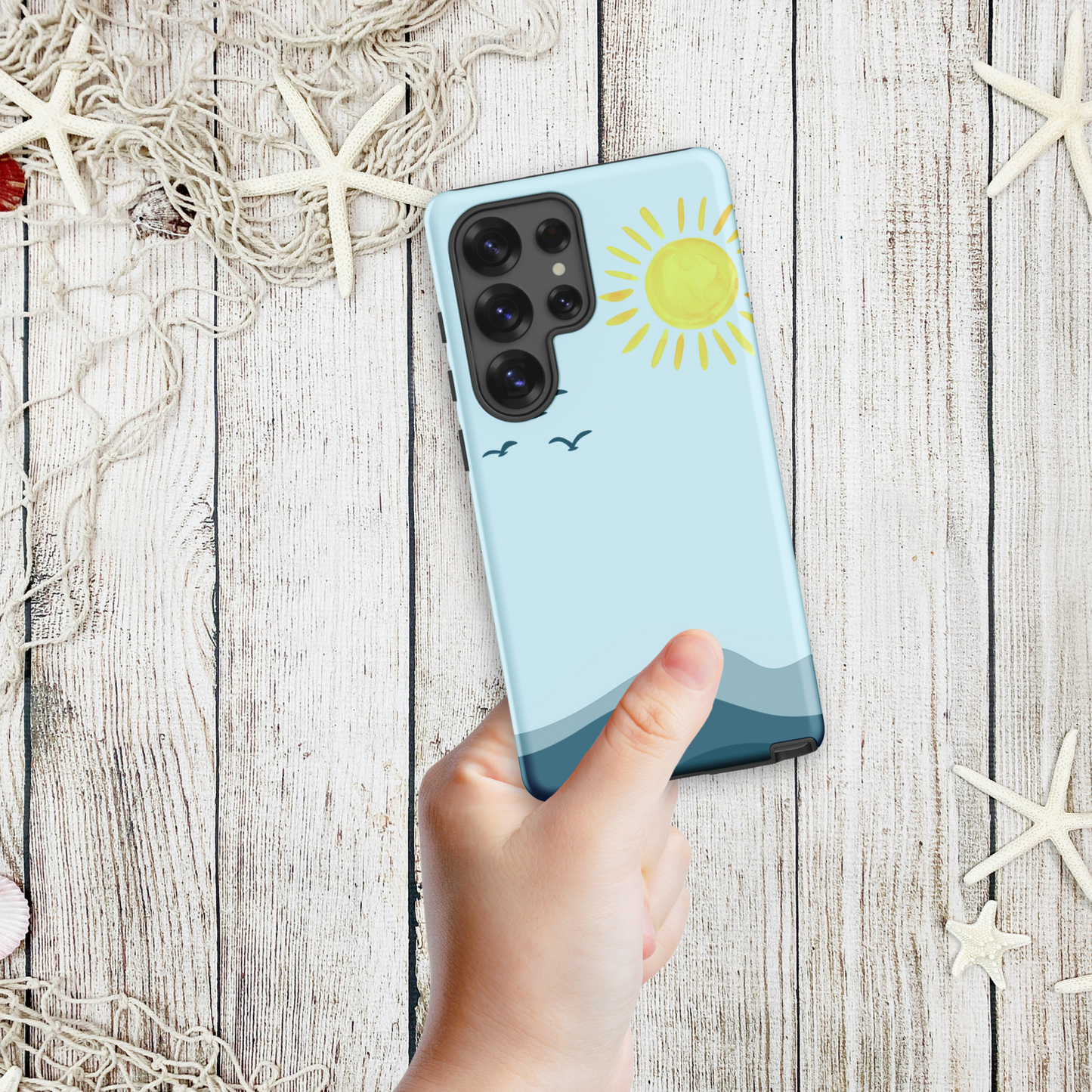 Hand holding a coastal Samsung phone case with a minimalist ocean horizon, yellow sun, and flying seabirds, styled on a weathered wood background with starfish.