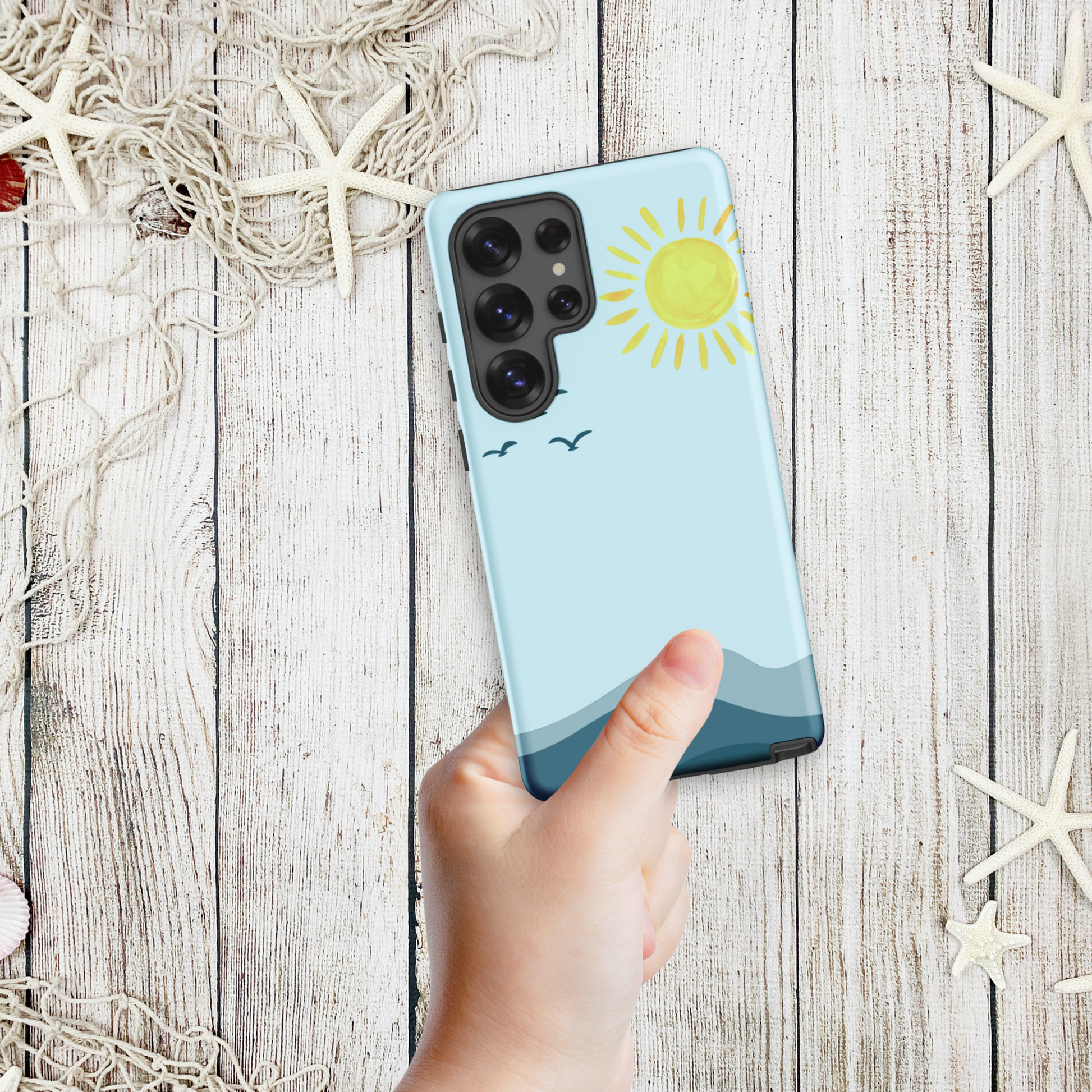 Hand holding a coastal Samsung phone case with a minimalist ocean horizon, yellow sun, and flying seabirds, styled on a weathered wood background with starfish.