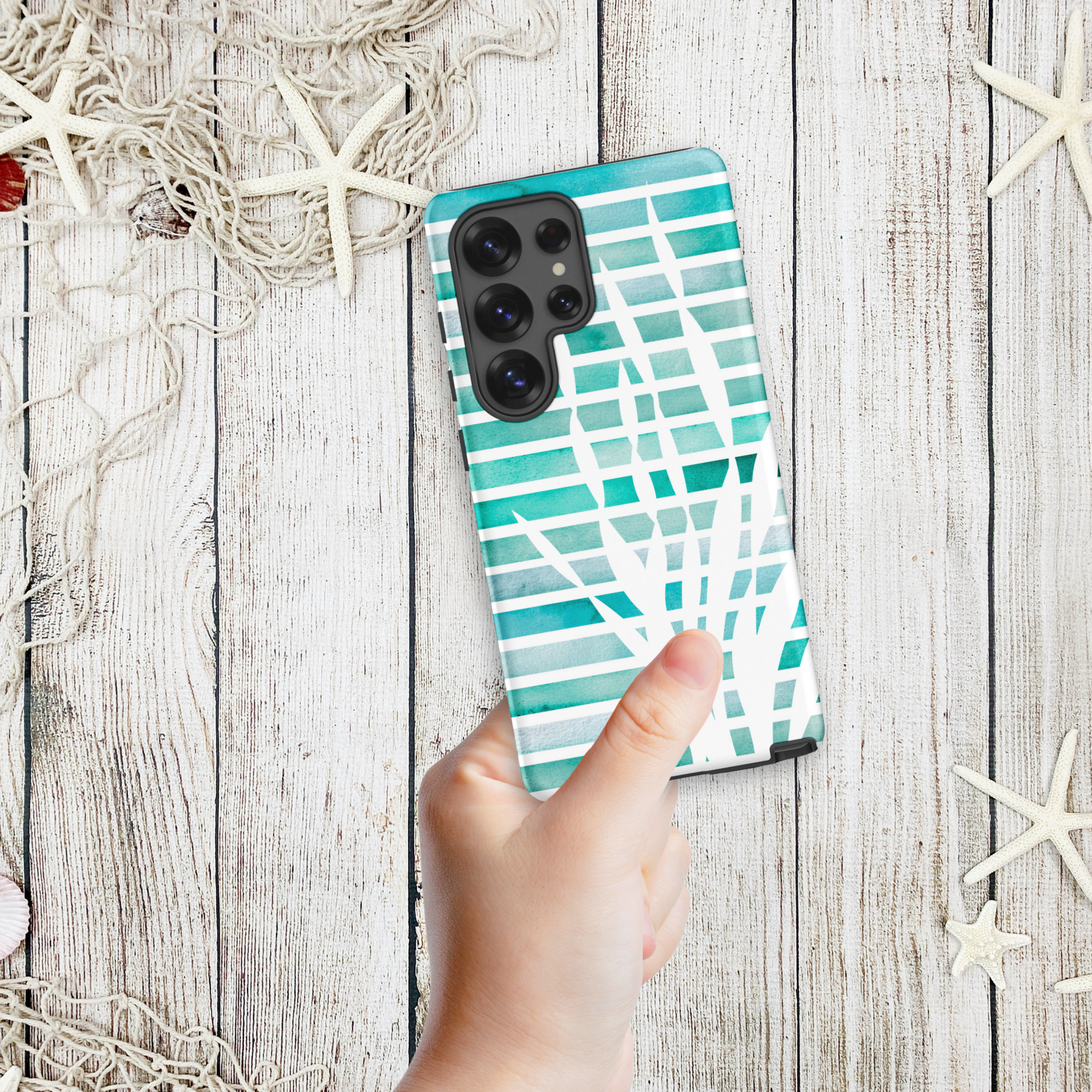 Hand holding a coastal Samsung phone case with abstract teal kelp fronds layered over watercolor stripes, styled with a nautical net and starfish.