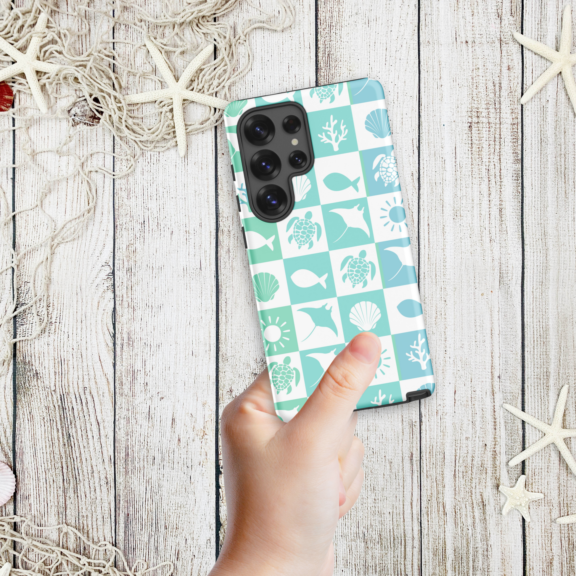 Hand holding a coastal Samsung phone case with aqua and white sea mosaic squares featuring shells, turtles, coral, and fish, styled on a wooden background with starfish.