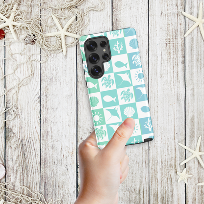 Hand holding a coastal Samsung phone case with aqua and white sea mosaic squares featuring shells, turtles, coral, and fish, styled on a wooden background with starfish.