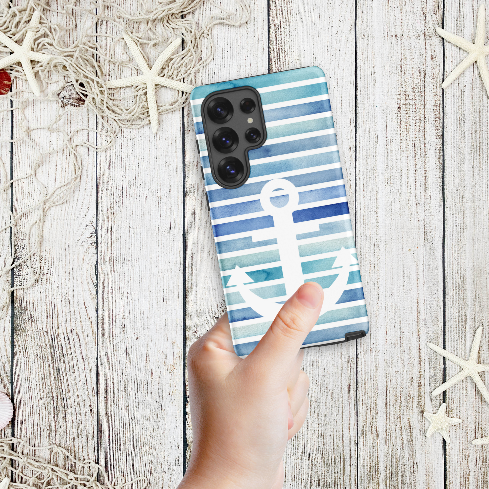 Hand holding a coastal Samsung phone case with navy and aqua stripes and a large white anchor, styled on a wood background with nautical netting and starfish.